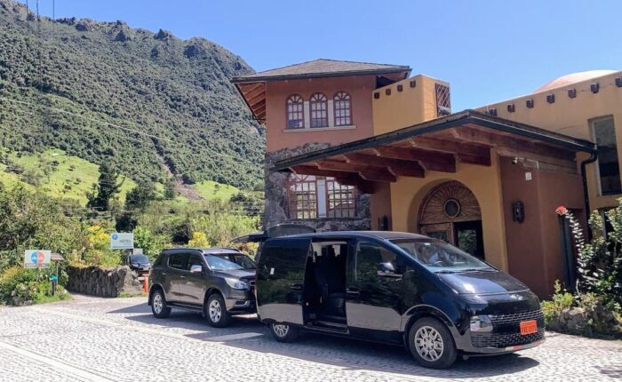 Black van with its sliding door open parked in front of a warm mountain hotel on a sunny day with green hills behind.