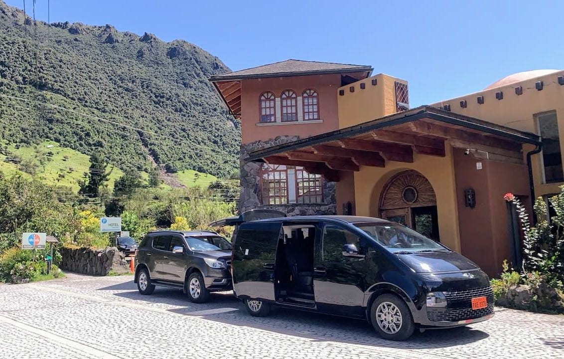 Black van with its sliding door open parked in front of a warm mountain hotel on a sunny day with green hills behind.